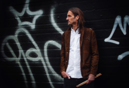 Dan Stefik satnds with drumsticks in hand in front of a black wall painted with graffiti