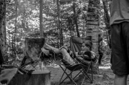 A boy lounges on a camping chair near a fire
